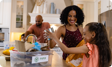 A smiling woman and young girl recycle cans in a kitchen, while a man sorts items in the background. A box labeled PAPER sits on the counter with recyclable materials inside.
