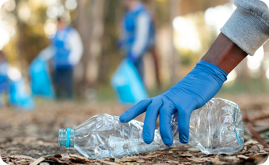 A person wearing a blue glove picks up a plastic bottle from the ground, with other people cleaning up litter in the blurred background outdoors.