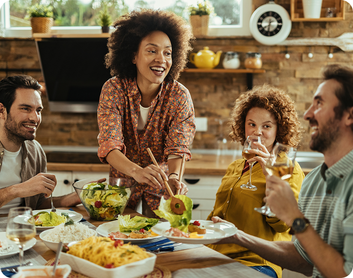 Four people sit around a kitchen table, smiling and enjoying a meal together. One woman serves salad, while others hold drinks and engage in cheerful conversation. The kitchen setting is warm and inviting.