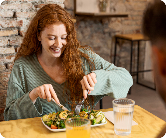A woman with long red hair smiles while cutting food on her plate at a restaurant table. She is wearing a light green sweater, and there are glasses of water and orange juice on the table.